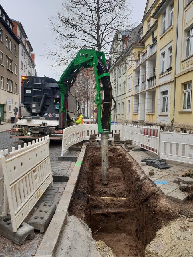 Bagger gräbt Löscher für Baumpflanzung an einem Straßenrand, Bauzäune begrenzen die Baugrube